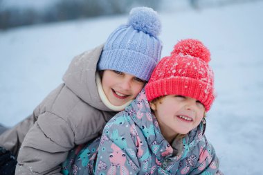 İki çocuk renkli şapkalar ve ceketler giyerek karda birlikte oynuyorlar. Etraflarında kar yağarken neşeli bir anı paylaşıyorlar, eğlenceli bir sahne yaratıyorlar..