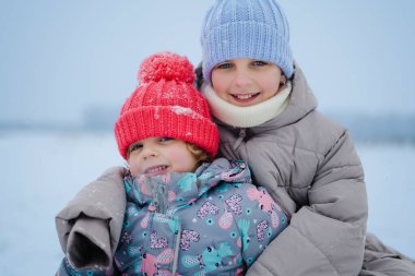 İki genç kız, parlak şapkalar ve kalın paltolar giyerek karda birlikte oturuyorlar. Kış manzarasıyla çevriliyken kameraya gülümserler..
