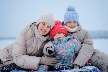 Bir anne iki çocuğuyla karla kaplı zeminde oturur. Kışlık giysiler giymişler ve kameraya gülümsüyorlar..