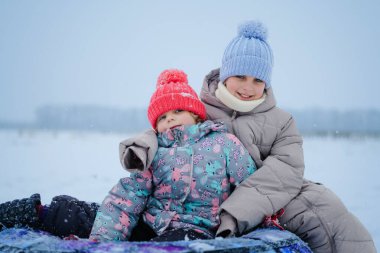 Soğuk bir günde, hafif kar yağışı olan neşeli bir anın tadını çıkarırlarken, iki çocuk karda kızağın üzerinde, kalın kışlık giysiler giyerek birlikte oturuyorlar..