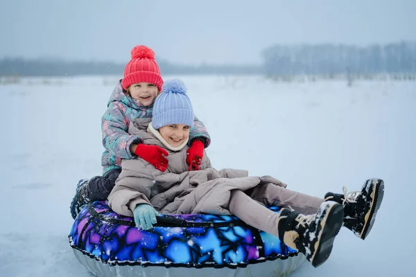 Karlı bir bölgede iki çocuk bir kar tüpünün üzerinde oturuyor. Gülümsüyorlar ve karda eğlenirken kışlık giysiler ve şapkalar giyiyorlar..