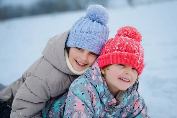 İki çocuk renkli şapkalar ve ceketler giyerek karda birlikte oynuyorlar. Etraflarında kar yağarken neşeli bir anı paylaşıyorlar, eğlenceli bir sahne yaratıyorlar..
