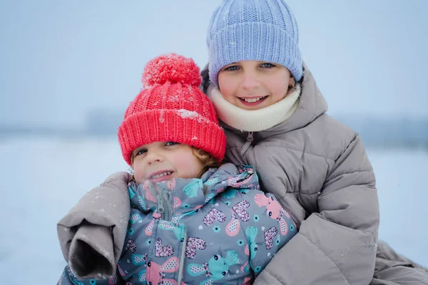 İki genç kız, parlak şapkalar ve kalın paltolar giyerek karda birlikte oturuyorlar. Kış manzarasıyla çevriliyken kameraya gülümserler..