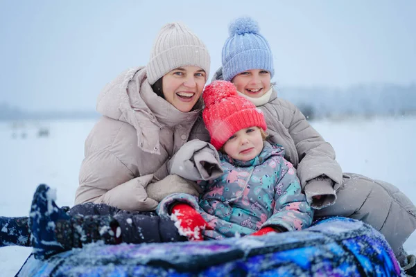 Üç kişi karda renkli bir kızağın üzerinde oturuyor, gülümsüyor ve birlikte vakit geçiriyorlar. Çocuklar karlı bir arazide oynarken sıcak şapkalar takarlar..