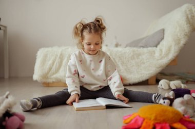 Bir kız önünde açık bir kitapla yerde oturuyor. Mutlu görünüyor ve aydınlık, güneşli bir odada oyuncaklarla çevrili..