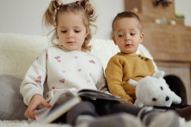 İki çocuk kanepede oturup bir kitaba bakıyor. Bir çocuk oyuncağını tutarken diğeri okur. Gün boyunca parlak bir oturma odasındalar..