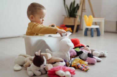 Bir çocuk beyaz bir kutuda oturur ve oyuncak bir arabayla oynar. Doldurulmuş hayvanlar ve oyuncaklarla çevrili güneşli bir odada..