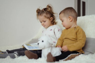 İki küçük çocuk sıcacık bir yüzeyde oturuyor. Bir çocuk kitap okurken, diğeri oyuncak bir köpek tutuyor. Hareketlerine sessizce odaklanırlar..