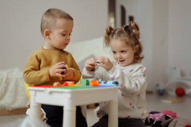 İki genç çocuk renkli yapı taşlarıyla bir masada oturur ve güneşli bir günde sıcak bir mekanda heyecan gösterirler.