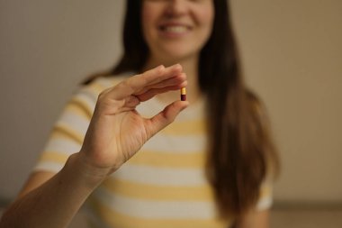 Bir kadın parmaklarının arasında küçük bir hap tutarken içeride oturuyor ve gülümsüyor. Doğal ışık, o haplara odaklanırken odayı dolduruyor..