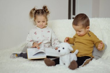 Biri kitap okurken, diğeri oyuncak bir köpekle oynarken iki çocuk yumuşak bir halıya oturur. Sakin bir öğleden sonra evde vakit geçirmeyi severler..