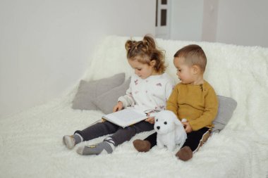 İki küçük çocuk beyaz bir kanepede oturup kitap okuyorlar. Yanlarına doldurulmuş bir hayvan konur. Onlar kitaptaki hikayeye odaklandılar..