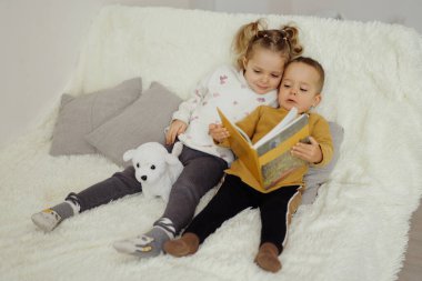 İki çocuk bir kanepede oturup kitap paylaşıyor. Yanlarında küçük doldurulmuş bir hayvan yatıyor. Oturma odasında kitap okumayı ve birlikte vakit geçirmeyi seviyorlar..