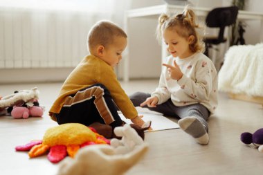 İki çocuk iyi ışıklandırılmış bir odada renkli oyuncaklarla çevriliyken yere oturup kağıda resim çiziyor. Birlikte oynarken düşüncelerini ve gülümsemelerini paylaşıyorlar..