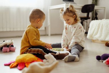 İki çocuk yerde oturup birlikte kitap okuyor. Odanın tadını çıkarırlarken pelüş oyuncaklar etrafa saçılmış..