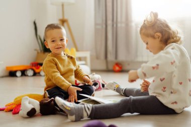 İki çocuk, doğal ışığı olan bir odada yerde oturuyor. Bir çocuk gülümserken diğeri etrafa dağılmış bir kitapla ve oyuncaklarla meşgul oluyor..