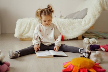 Bir çocuk önünde açık bir kitapla yerde oturuyor. Parlak bir odada oyuncaklar etrafa saçılırken sayfaları işaret ediyor..