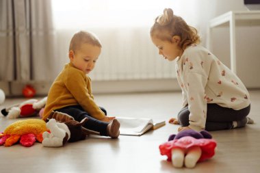 İki çocuk yerde oturup kitap okurken oyuncaklar etrafa saçılmış. Güneş ışığı odaya girer ve sıcak bir atmosfer yaratır..