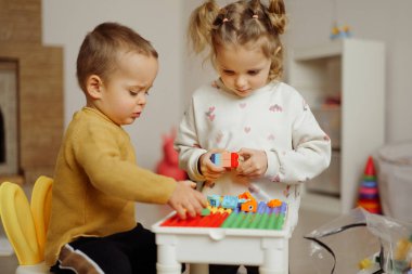 İki küçük çocuk küçük bir masada birlikte oynuyorlar. Parlak bir oyun odasında renkli blokları olan yapılar inşa ediyorlar..