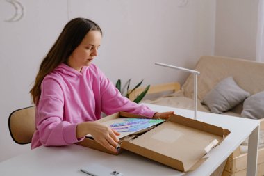 Genç bir insan parlak bir odada bir masada oturuyor, renkli bir sanat içeren bir kutuyu açıyor. Bir lamba mekanı aydınlatıyor ve yakınlarda bir kanepe var..