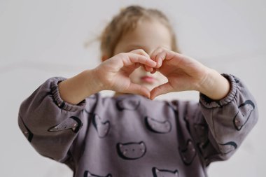 Parlak bir odada duran bir çocuk elleriyle kalp şekli oluşturuyor. Çocuk sevgi ve neşe ifade ederken odaklanmış bir ifadeye sahiptir..