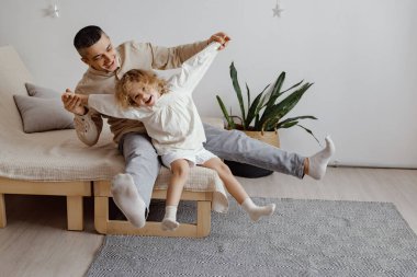 Baba çocuğunu oturma odasında sallıyor. Açık renkli bir kanepede oturuyorlar ve yakında bitkiler var. Sıcak öğleden sonra güneşi pencereden parlıyor..