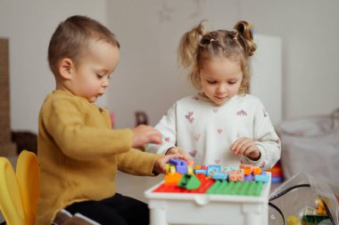 İki çocuk bir masada oturup renkli yapı taşlarıyla oynuyorlar. Kapalı bir alanda farklı şekiller ve yapılar inşa etmeye odaklanırlar..
