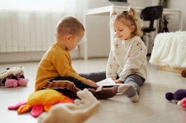İki çocuk aralarında bir kitapla yerde oturuyor. Güneşli bir odada yumuşak oyuncaklarla çevrili rahat bir atmosferi ve yakınlarda mobilyaları var..