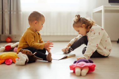 İki çocuk aydınlık bir zeminde oturup bir kitaba bakıyor. Etraflarında oyuncaklarla oynuyorlar. Pencereden içeri güneş ışığı giriyor, odayı aydınlatıyor..