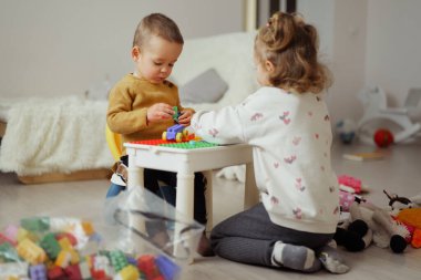 İki çocuk bir masada renkli yapı taşlarıyla oturma odasında oynuyorlar. Oyuncaklar etrafa saçılırken onlar aktivitelerine odaklanırlar..