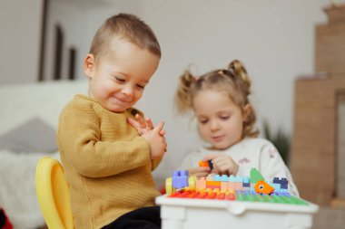İki çocuk yerde oturuyorlar. Bir çocuk oyuncağa odaklanırken diğer çocuk izliyor ve oynuyor. Parlak bir kapalı alandalar..