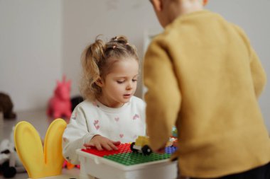İki çocuk bir masada renkli yapı taşlarıyla oynuyorlar. Odaklanıyorlar ve yumuşak ışığın olduğu bir odada birlikte vakit geçiriyorlar..