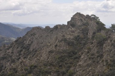 Dik yamaçlı kayalık dağ sırtı, seyrek çam ağaçları ve Kıbrıs 'ta bulutlu gökyüzü altında katmanlı Akdeniz manzarası
