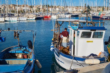 İtalya, Sicilya, Marina di Ragusa (Ragusa Eyaleti); limanda ahşap bir balıkçı teknesi ve lüks yatlarda çalışan balıkçı. 