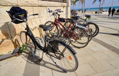 İtalya, Sicilya, Akdeniz, Marina di Ragusa (Ragusa Eyaleti), 27 Şubat 2021, parketmiş bisikletler ve palmiye ağaçlarıyla deniz kenarında yürüyen insanlar