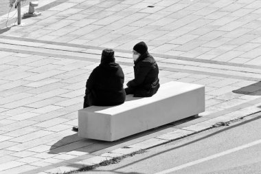 İtalya, Sicilya, Akdeniz, Marina di Ragusa (Ragusa Eyaleti); limandaki bir bankta oturan cerrahi maskeli insanlar 