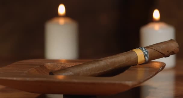 4K: Movimiento deslizante de un cigarro cubano en un cenicero con algunas velas en el fondo a una caja abierta de cigarros sobre una mesa de madera rústica . — Vídeos de Stock