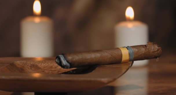 4K: Movimiento deslizante de un cigarro cubano brillante en un cenicero con algunas velas en el fondo a una caja abierta de cigarros en una mesa de madera rústica . — Vídeo de stock