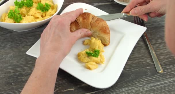 4K: Tagliare il croissant aperto e alcune uova strapazzate si trovano su un piatto bianco angolare. Una donna sta mettendo delle uova strapazzate fresche al croissant. . — Video Stock