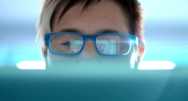 4K: A woman is using a tablet to play an online game which is reflected in her glasses. Her face is partly covered with a notebook. The background shows a blueish hightec enviroment with a lensflares — Stock Video