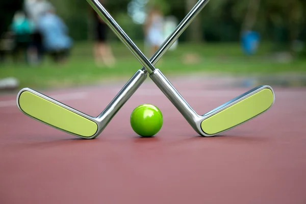 两个交叉的迷你高尔夫铁球拍和一个绿色的球在迷你高尔夫游乐场 — 图库照片