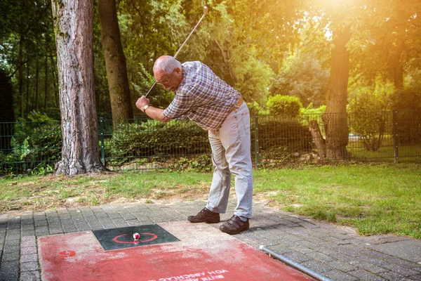 Old man at a minigolf court hits a ball with an iron racket. — Stock Photo, Image
