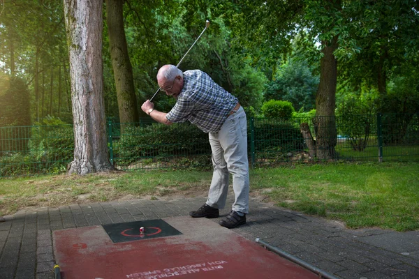 Male pensioner at a minigolf court hits a ball with an iron racket — Stock Photo, Image