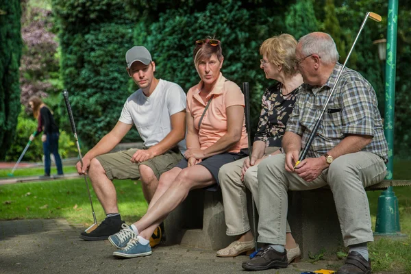 Two generation of peoples are sitting on a park bench at a minigolf court