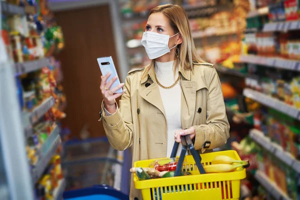 Mulher adulta em máscara médica usando smartphone e compras para mantimentos — Fotografia de Stock