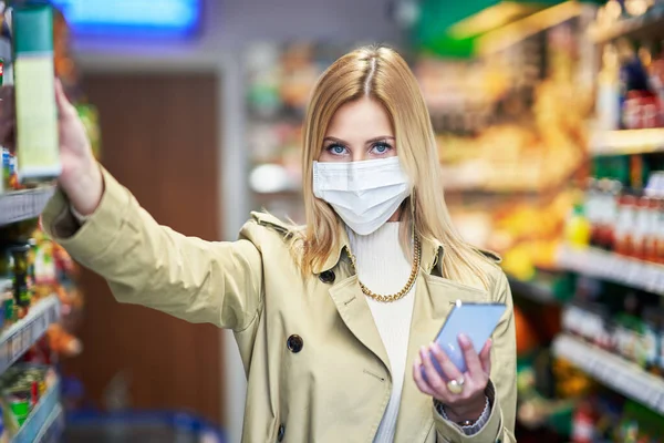 Mulher adulta em máscara médica usando smartphone e compras para mantimentos — Fotografia de Stock