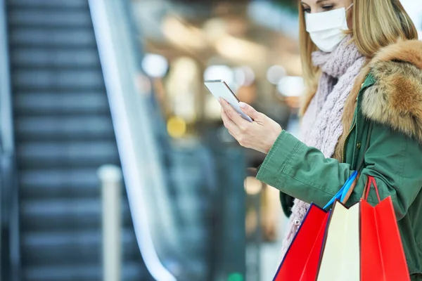 Middel av vuxen kvinna shopping i köpcentret med hjälp av smartphone bär mask — Stockfoto