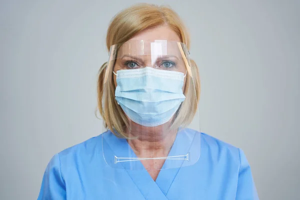 Médica sênior posando em máscara e escudo sobre fundo cinza — Fotografia de Stock
