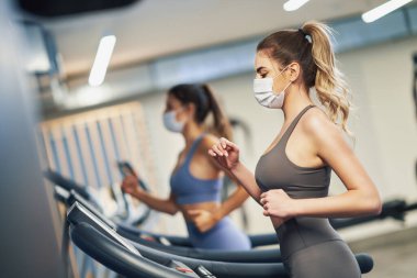 Maskeli iki kız arkadaş koşu bandında çalışıyor.