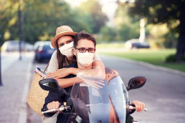 Güneşli bir günde şehirde scooter sürerken maske takan mutlu, olgun bir çift.